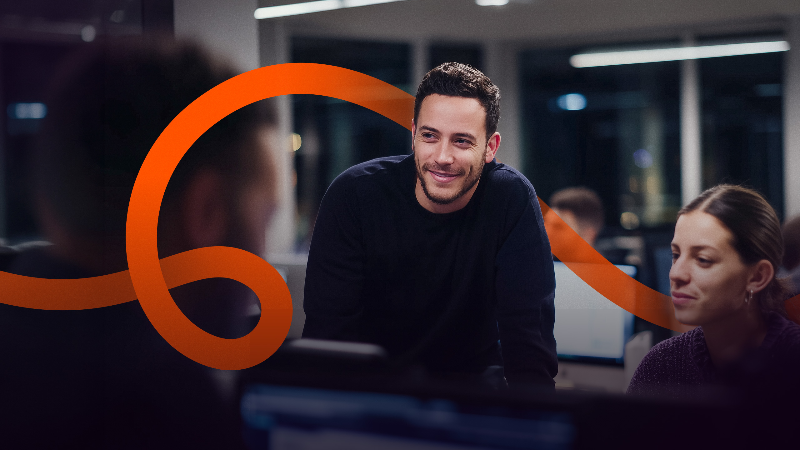 Man smiling in a night office as a colleague helps him at the monitors, with the orange line running across the frame
