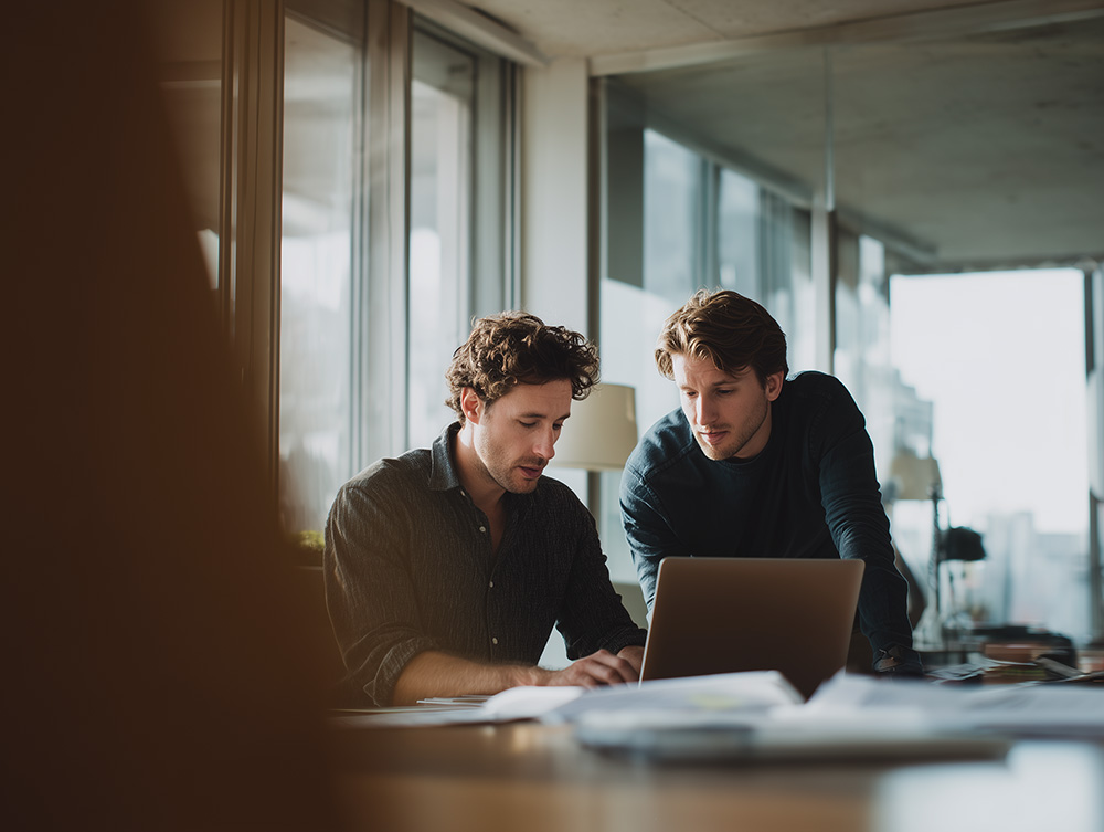 Two men collaborating over a laptop in a bright room lit by a window