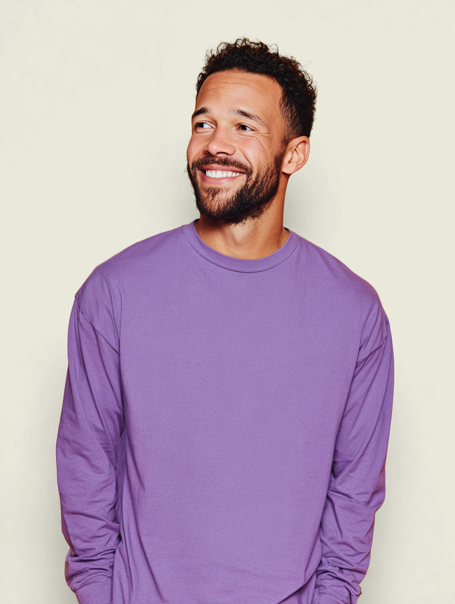 Editorial portrait of a man in a purple sweater smiling against a cream background, framed at eye level