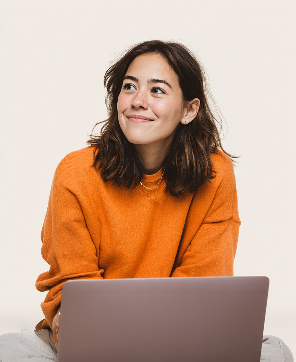 Woman in an orange sweater smiling while using a laptop against a cream studio background