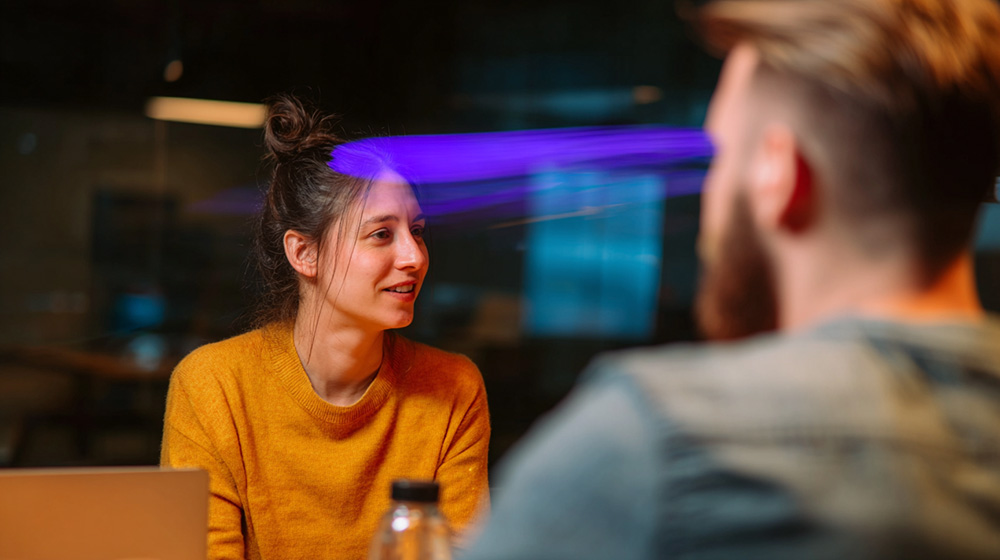 Woman in a mustard sweater talking with a colleague in an office with a purple light accent
