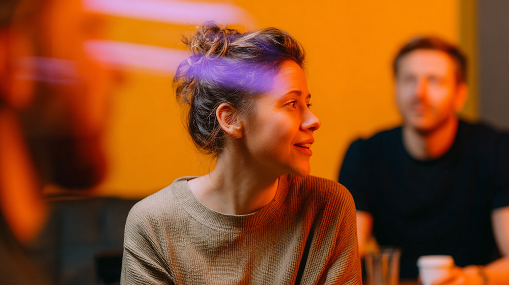 Woman in an olive sweater talking in a café with a yellow wall and a purple light accent