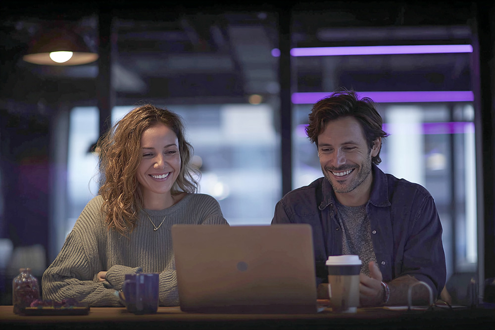Woman and man laughing while looking at a laptop in a night office with purple accents