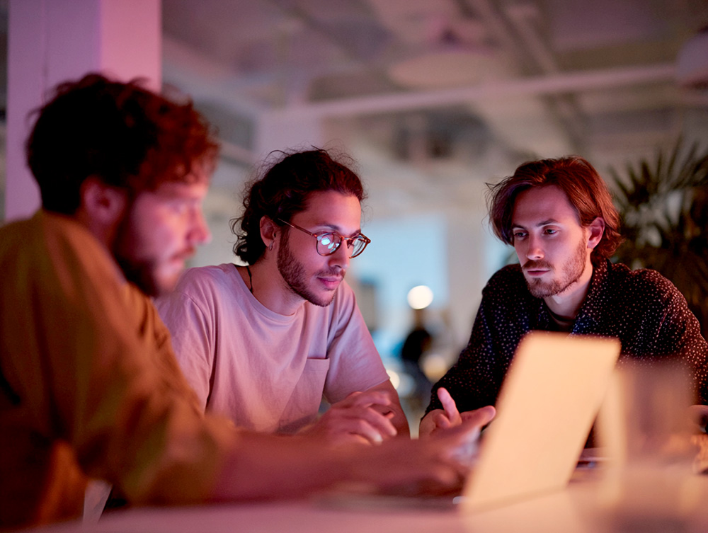 Three men focused around a laptop in an office under warm cinematic light